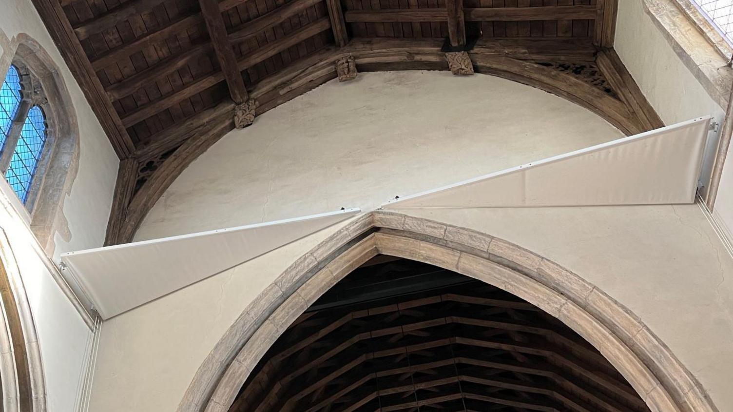 Two canvas sail-type structures under the roof of a church. They are held in place by fittings on a wall. The church roof is wooden. Arched windows can be seen too.