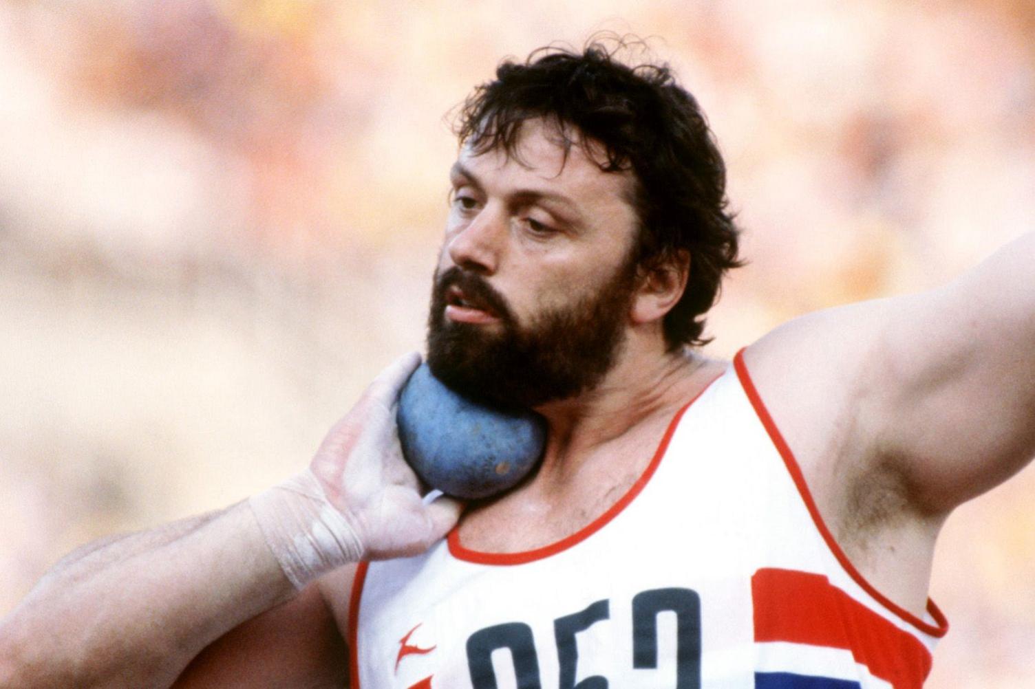 A picture of Geoff Capes competing in an outdoor event, holding a shot under his chin as he prepares to put it. He has a thick beard and is wearing a white-and-red athletics vest.