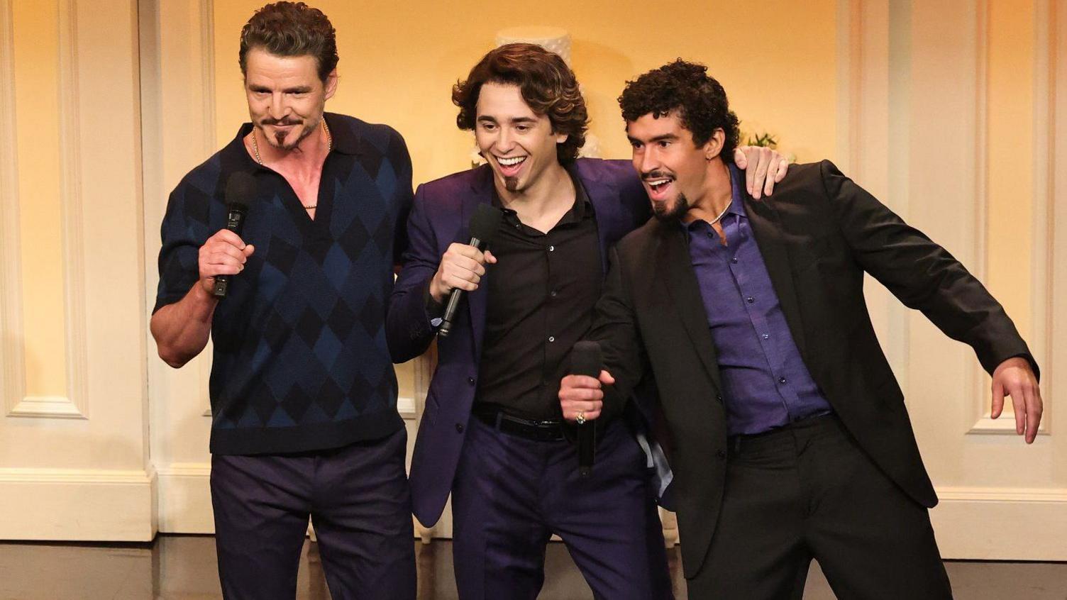 Three men stood together, with various facial hair. They all hold microphones, left one wears a diamond check short sleeved top, the middle wears a purple suit and the right one wears a black suit with a purple shirt