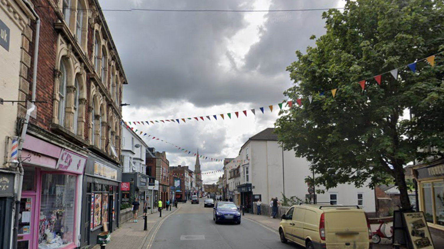 Fisherton Street in Salisbury, Wiltshire. Shops and pubs can be seen either side of the street. Cars can be seen travelling along the road whilst a van is parked up on the kerb. Bunting is hanging, stretching across the road. 