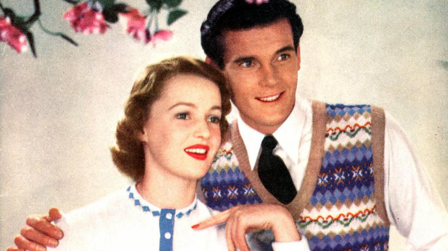 A young Sir Roger Moore is seen modelling knitwear. He is handsome with dark brown hair and a smile on his face. He is wearing a patterned tank top which is predominantly blue with diamond shapes on it, but it also features red and green love hearts. He has his arm around a woman who has brown curly hair and bright red lipstick on. She is wearing a white jumper with a blue decorative pattern around the neckline. Like Sir Roger, she is gazing into the distance.