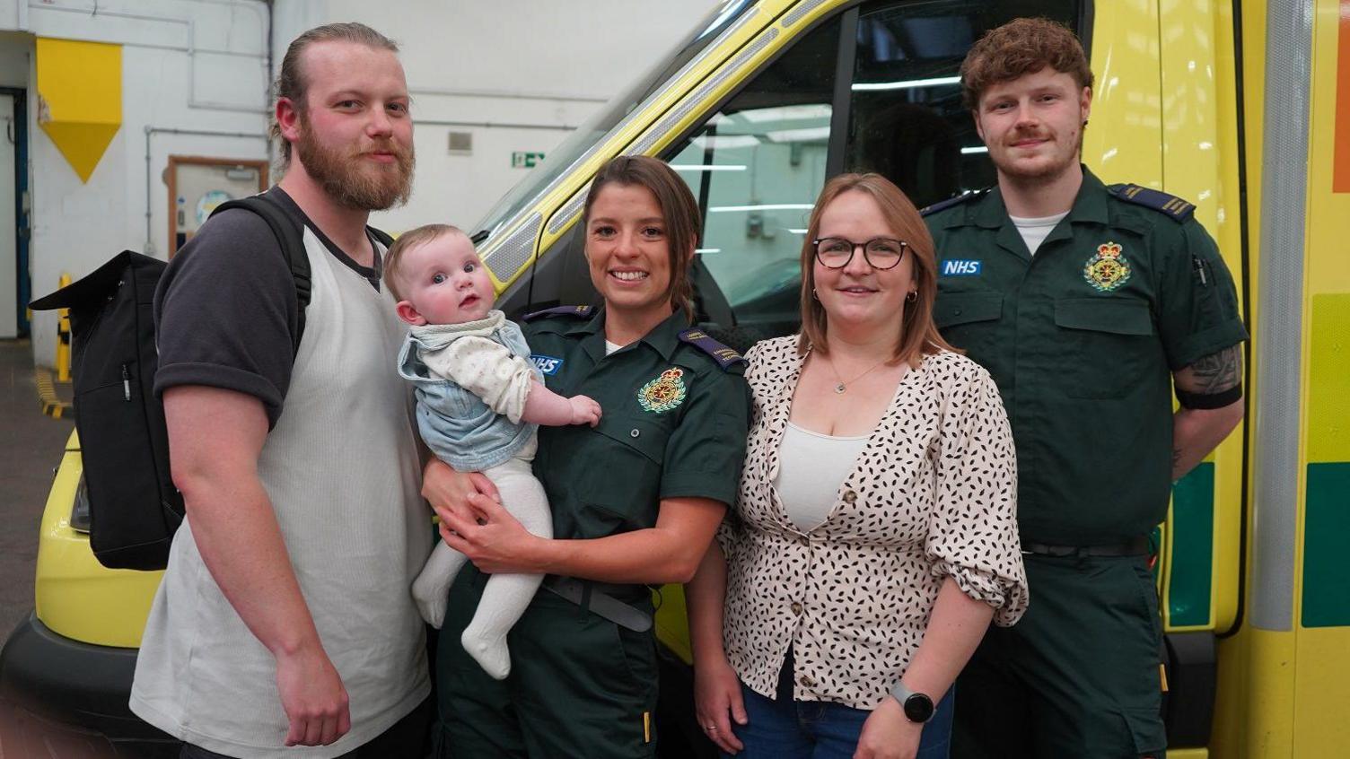 A group photo showing Dad Michael Everett who is wearing a grey t-shirt. Next to Michael is paramedic Courtney Mulligan who is wearing a paramedic uniform and holding baby Lydia Everett. Next to Courtney is mum Abie Everett wearing a blouse with black small spots on it. Next to Abie is another medic Corey Graham who is also wearing a medic's uniform and has his hands behind his back. All five are in front of an ambulance vehicle.