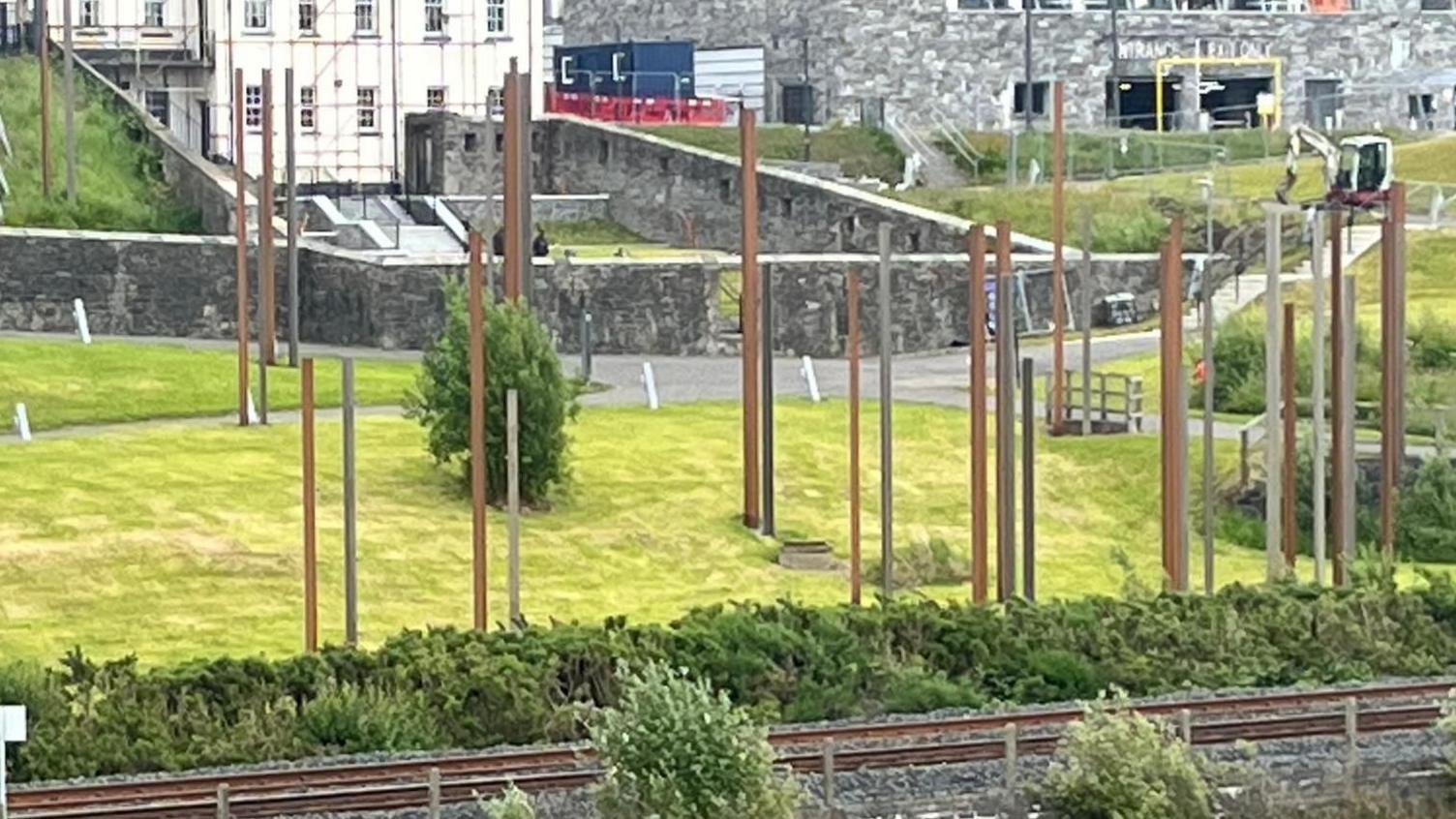 an array of steel pillars stand upright in an area of grass. a railway line is in the foreground