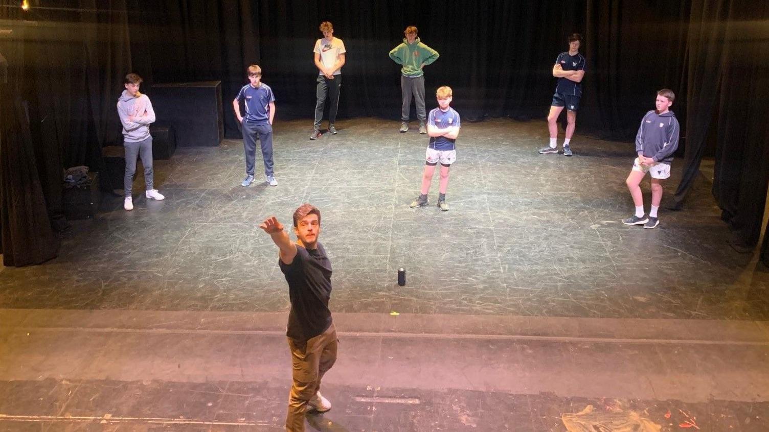 Jonno is on a stage at Bedford School gesturing. He is teaching a group of children stood behind him acting techniques.