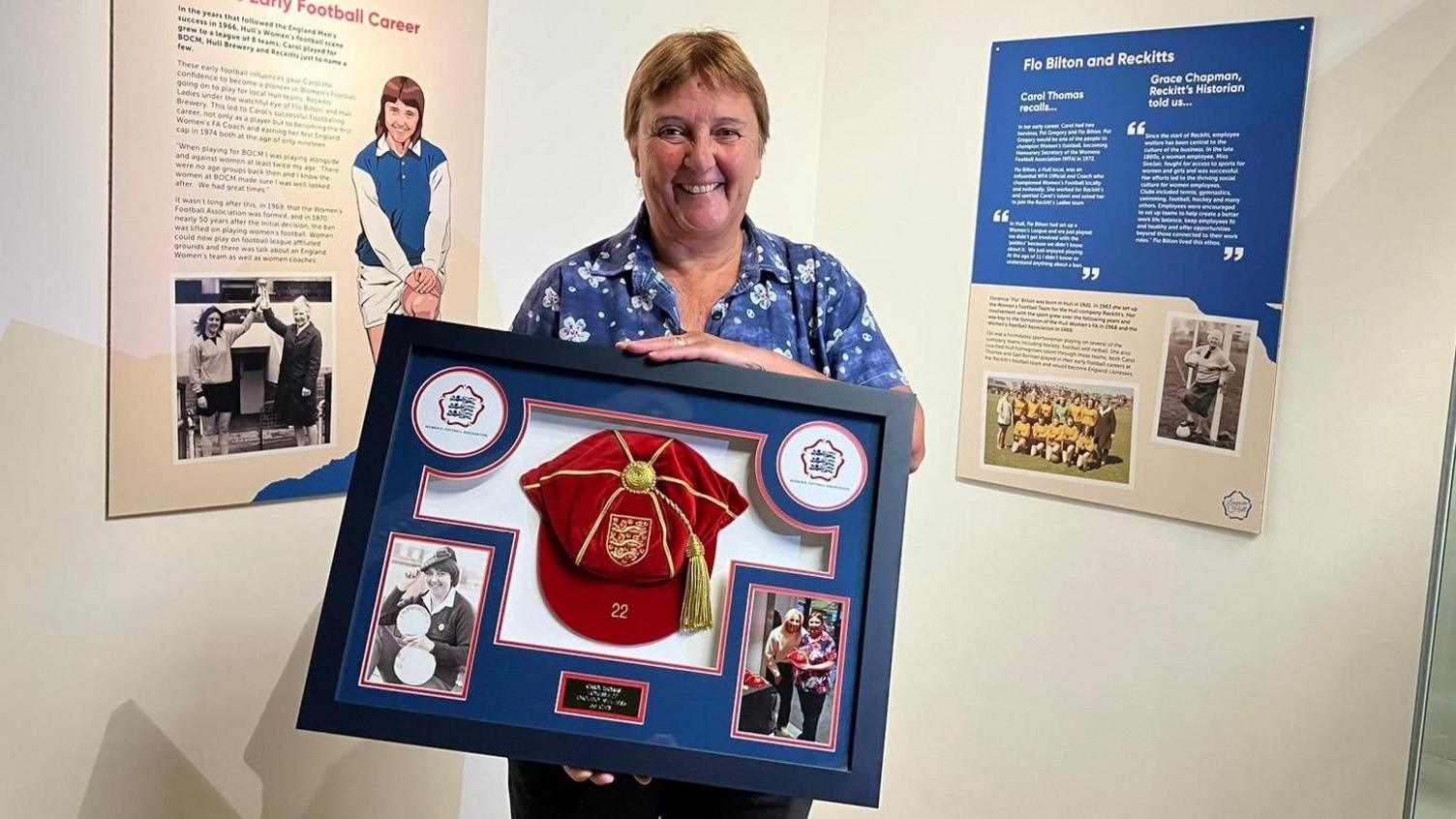 Carol Thomas, with short fair hair and a blue floral shirt, holds a museum display featuring a red and gold cap she was awarded for her footballing success. She stands between two display boards about her career at the Streetlife Museum's exhibition.