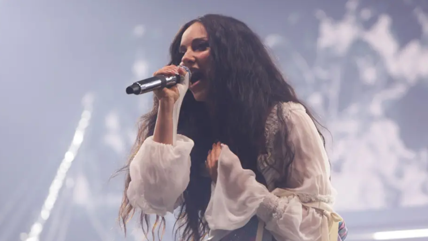 Jade Thirlwall is singing into a microphone while wearing a white blouse with baggy sleeves.