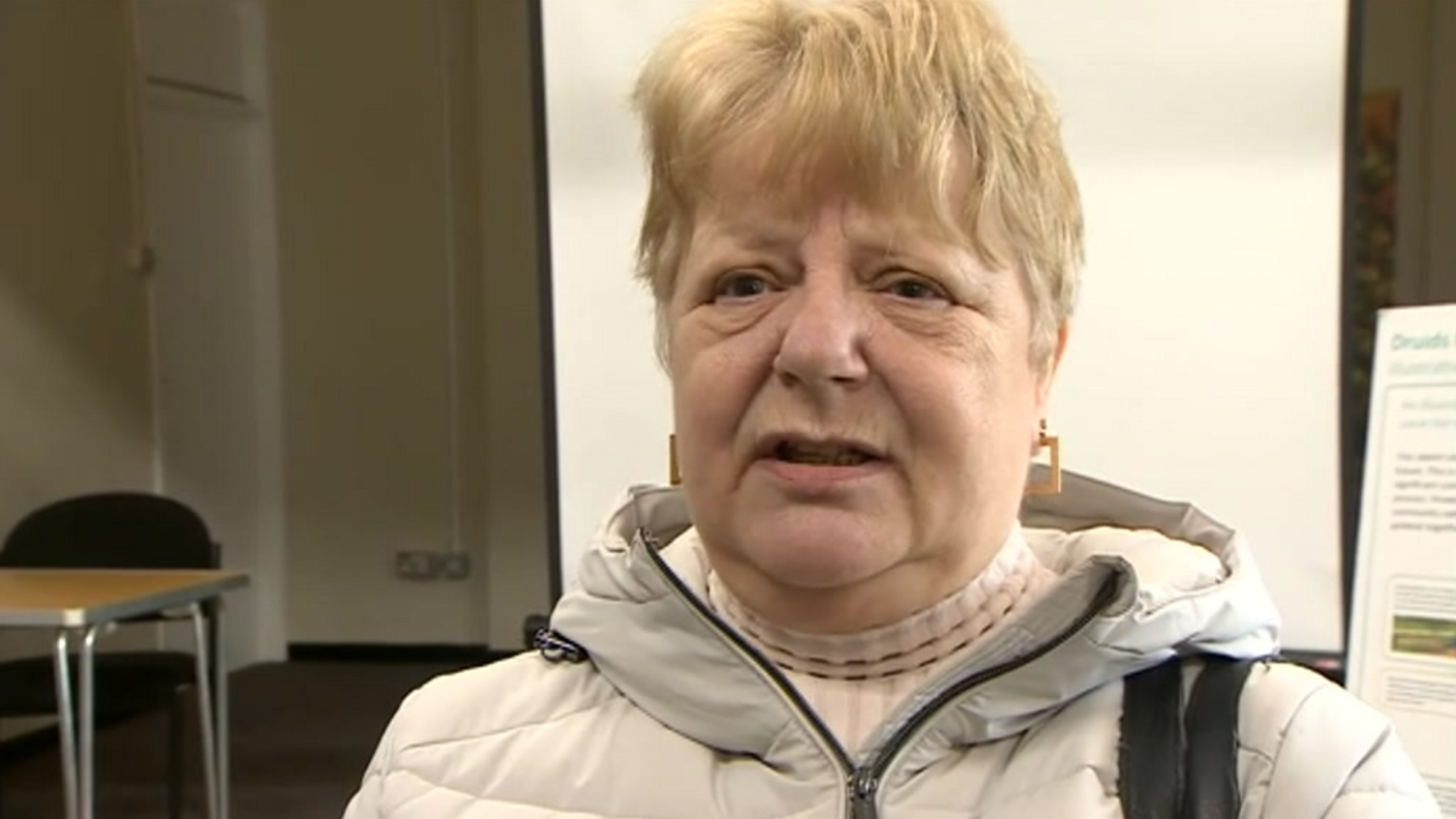 A woman with blonde hair and a white coat and jumper in a small room with a white screen behind her, as well as a chair and table