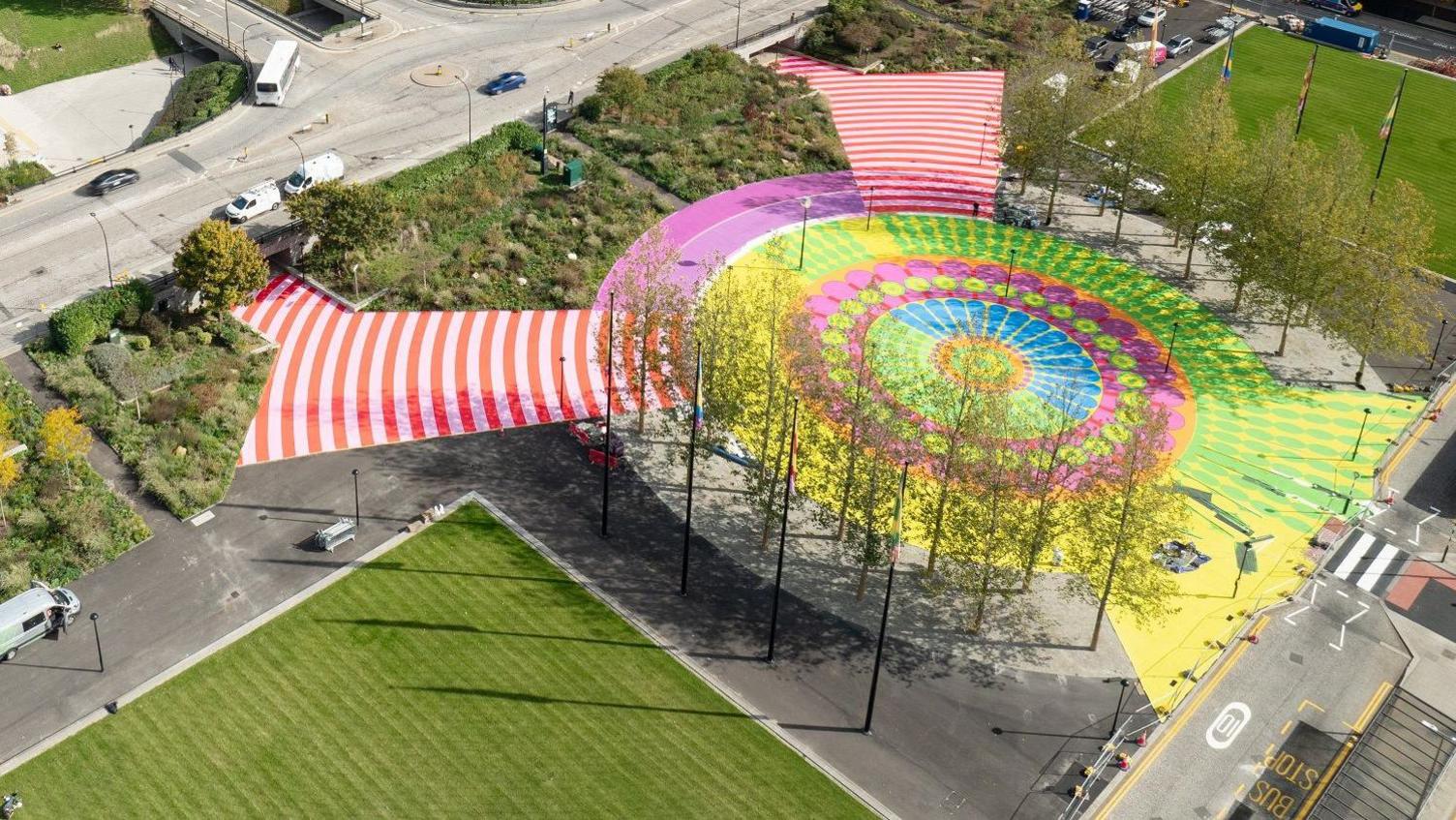 An aerial shot of Station Square with the new artwork. It is shaped in a circle that is edged with tall trees. Concentric circles show vivid patterns of yellow, lime green, fuschia pink and blue. Two red-and-white striped walkways lead out from the circle to two underpasses. Outside the circle are tall flagpoles with various flags, and on either side are large grass squares.