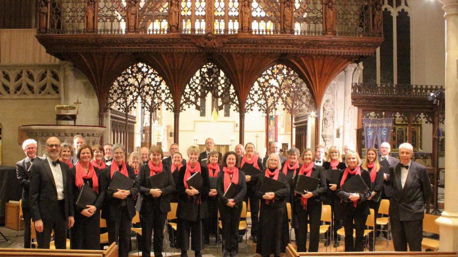 A large group called the Farrant Singers standing in a church all smiling at the camera. They are all dressed in black with red scarves. Some hold folders with their music in