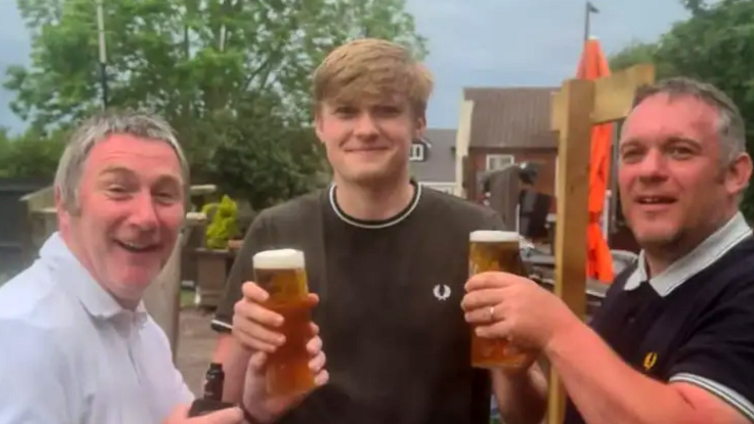 Three men in a line. The one in the middle is in his mid-20s, wearing a black top with white hems. Either side are two brothers in their 50s who look very similar, both with grey hair, wearing polo shirts and holding up pints.