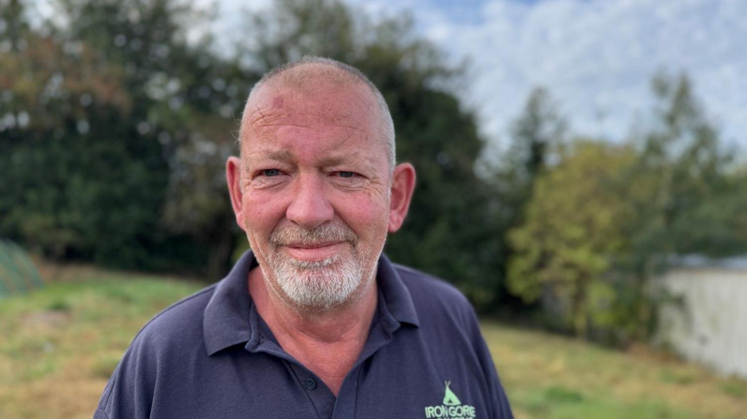 A man with a white beard is wearing a blue top. Grass behind him is blurred.