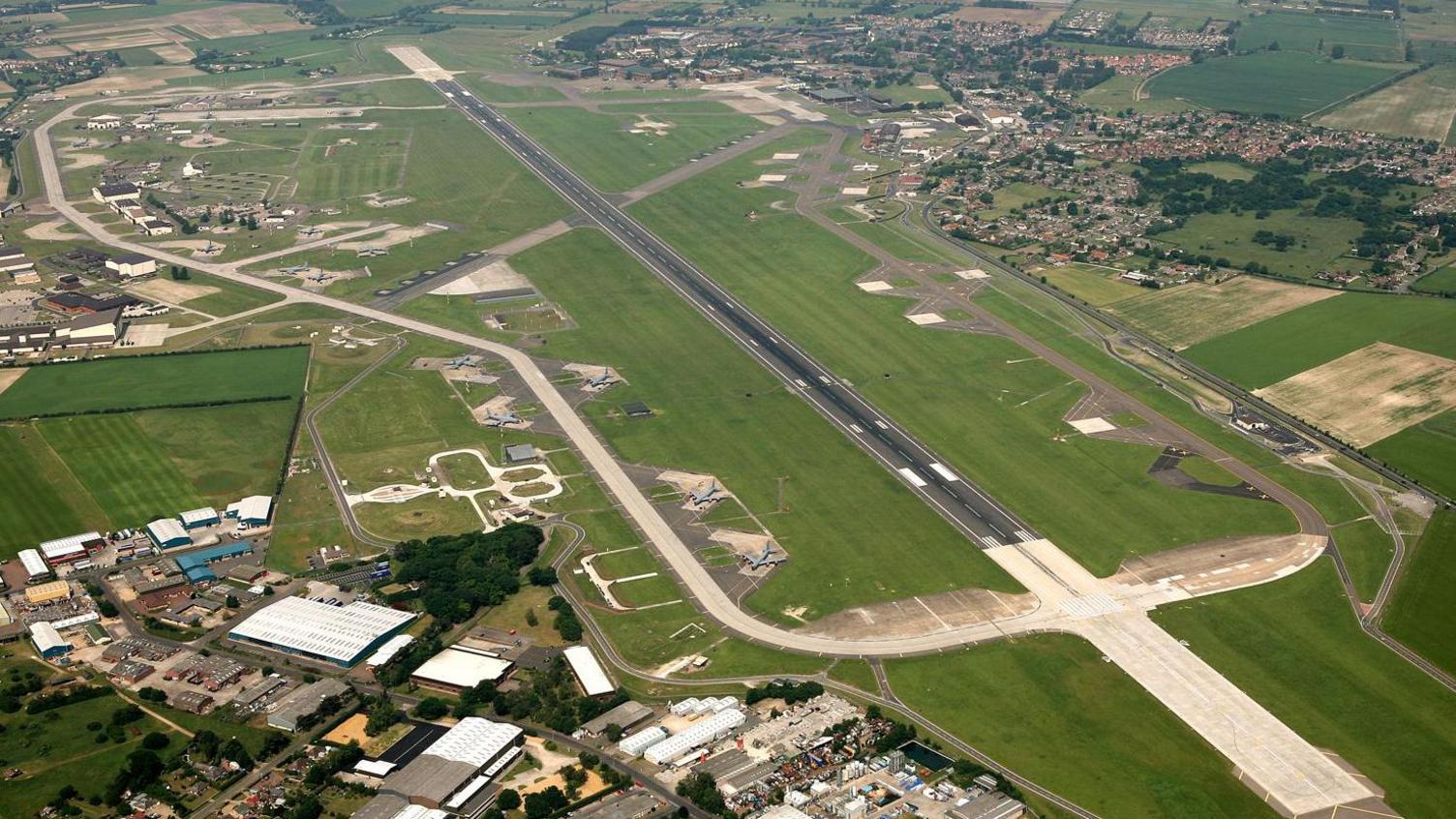 RAF Mildenhall airfield seen from the air, featuring a runway surrounded by grass and a perimeter road. An industrial estate is at the bottom of the picture, with the village of Beck Row at the top right.