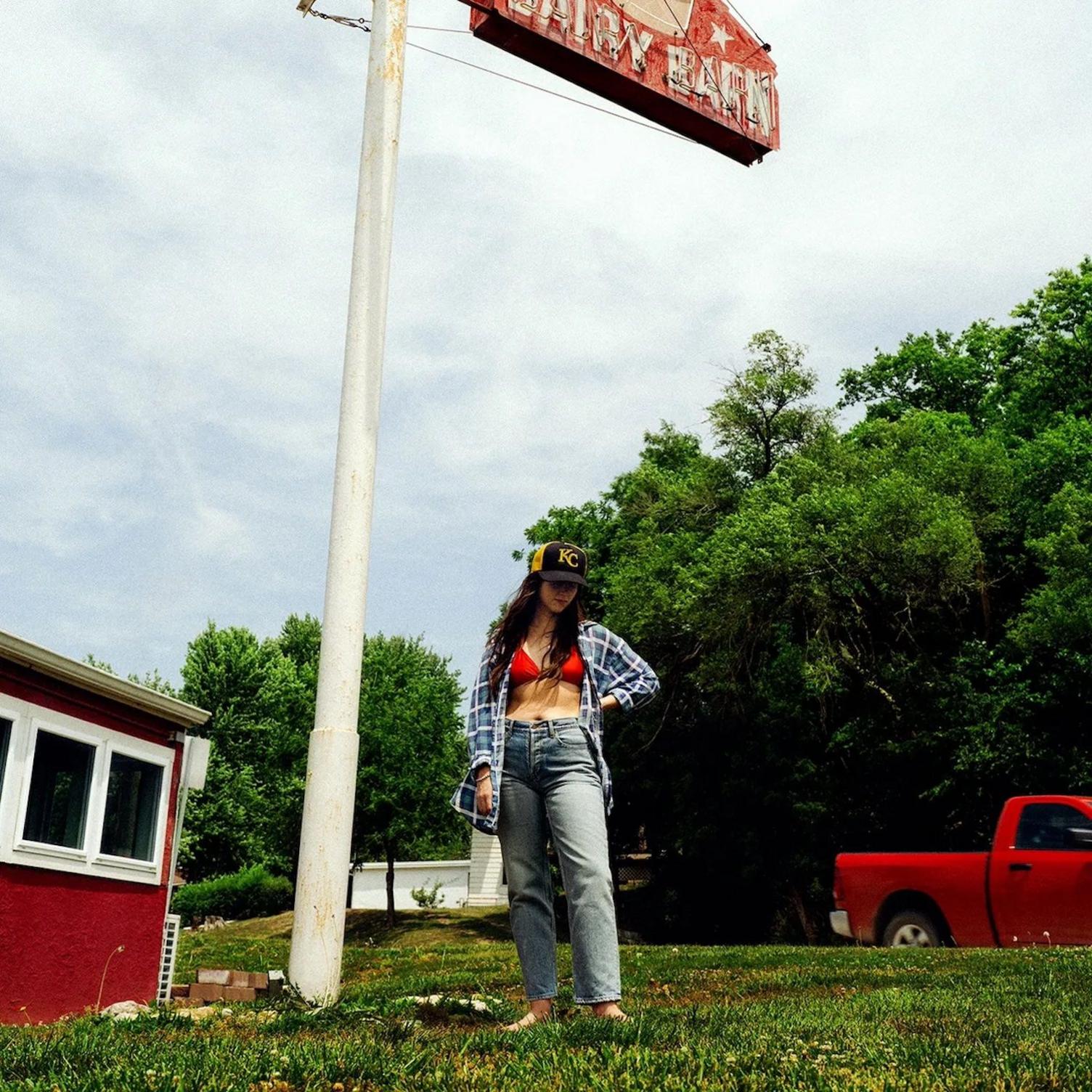 Artwork for Waxahatchee's album, Tigers Blood. A woman in jeans and an open  chequered shirt, which shows red bikini style top. She is standing on grass. In the background are trees, a red pick up and a small red building