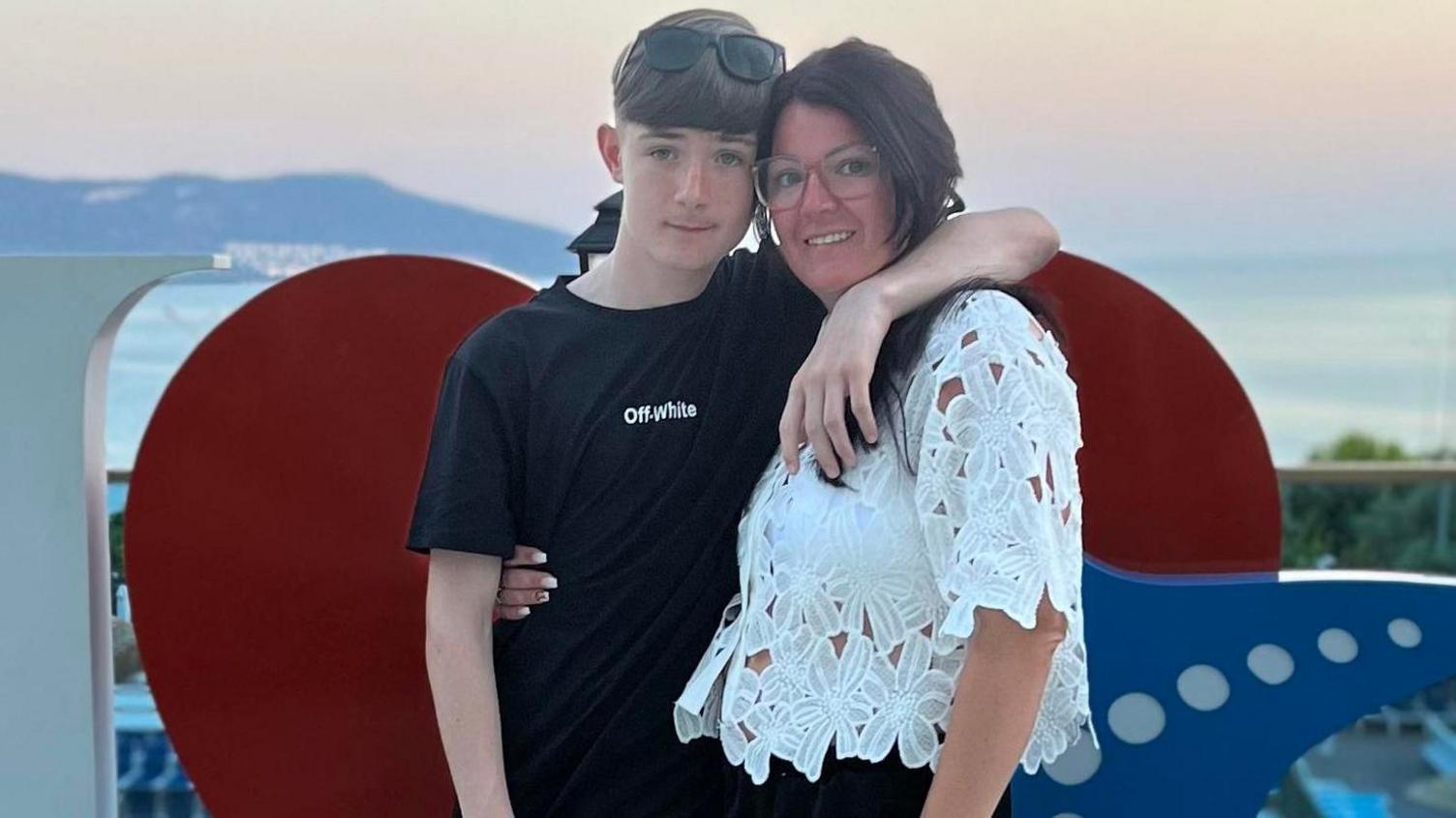 Carly Calland with long dark auburn hair and black glasses wearing a white top.
Her son has short hair, black t-shirt with white letters saying Off-white
