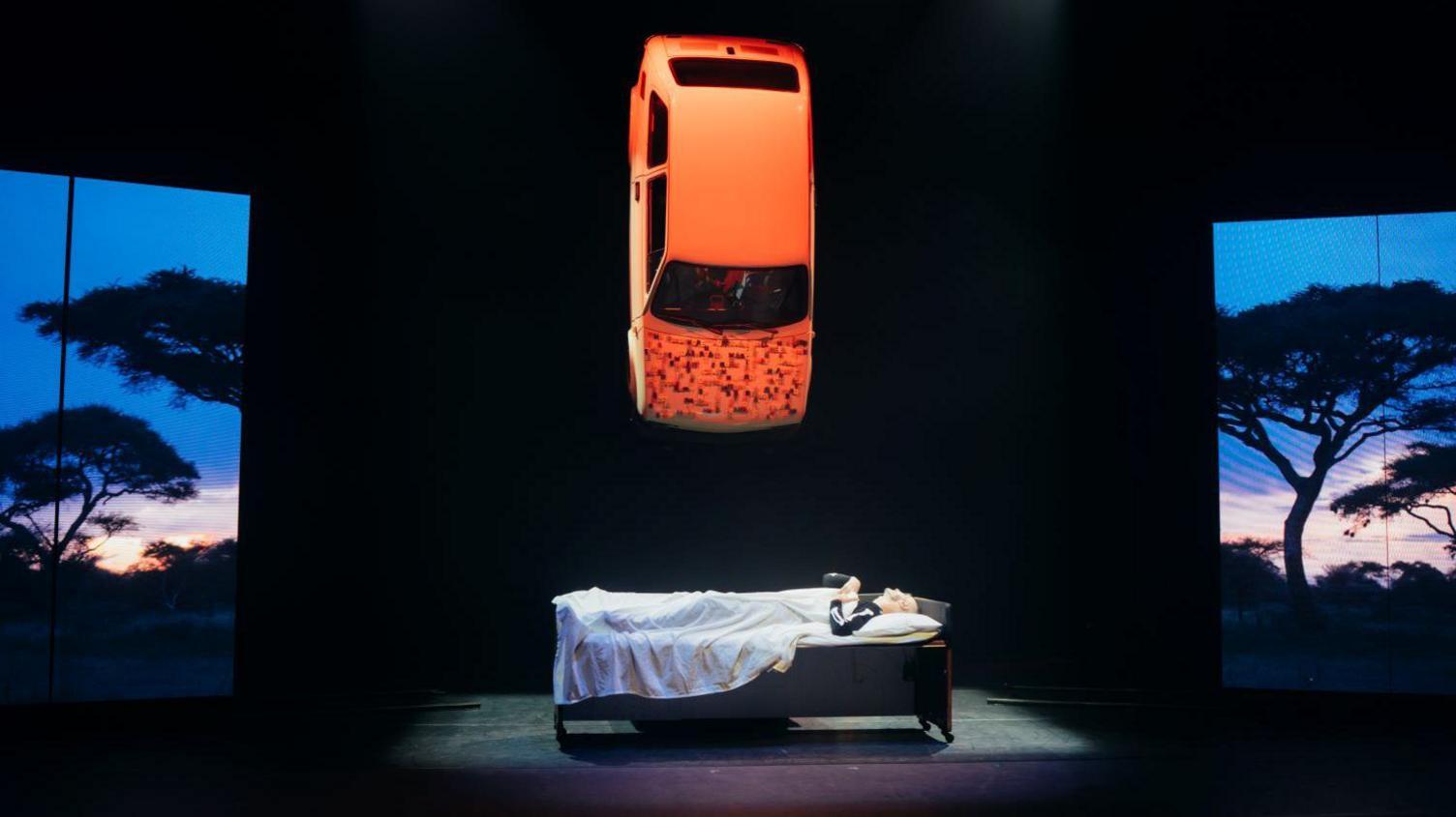 Marc Brew on stage lying in a bed with a car dangling above him