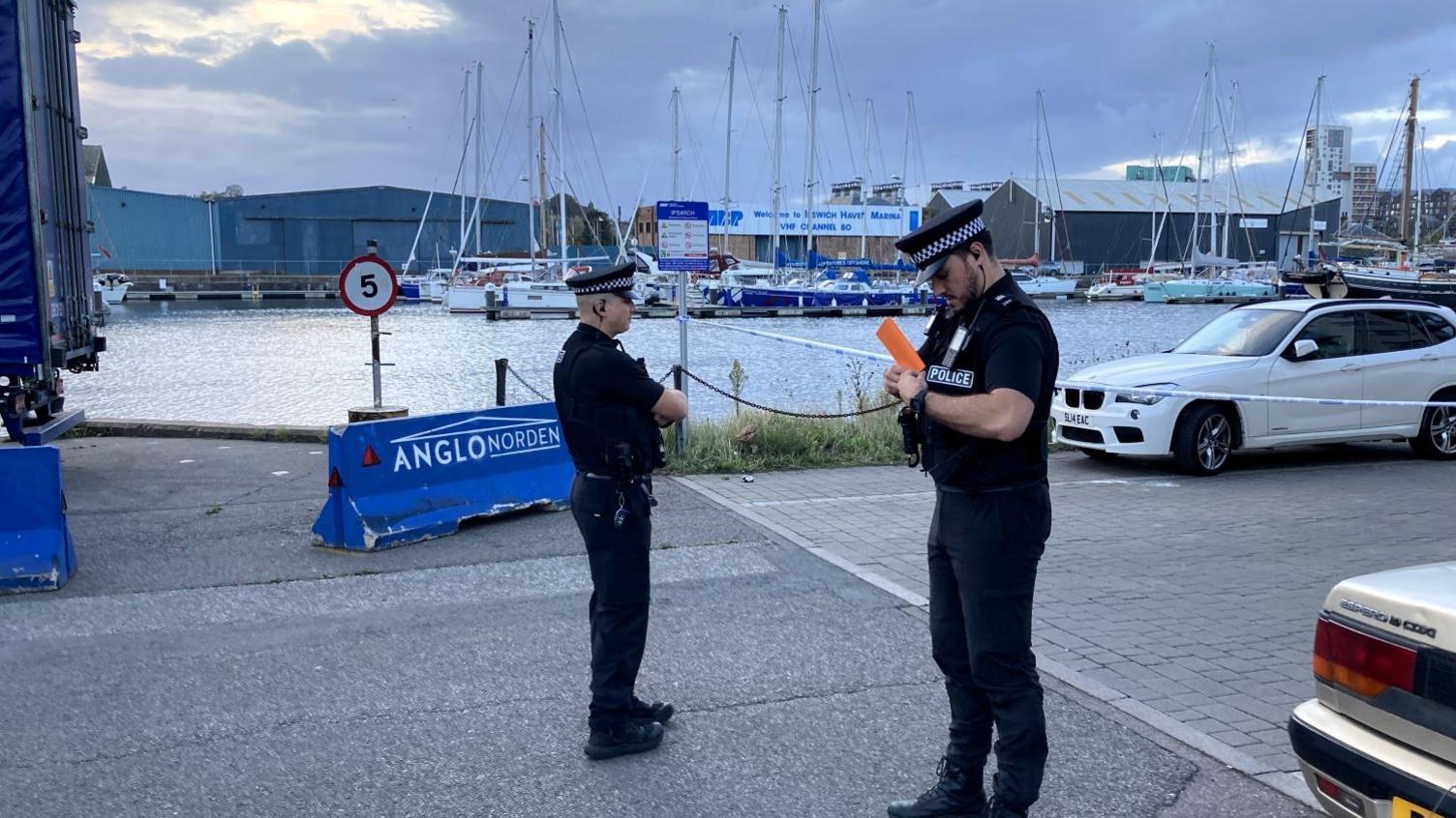 Two police officers stood by Ipswich Waterfront with boats and warehouses in the background