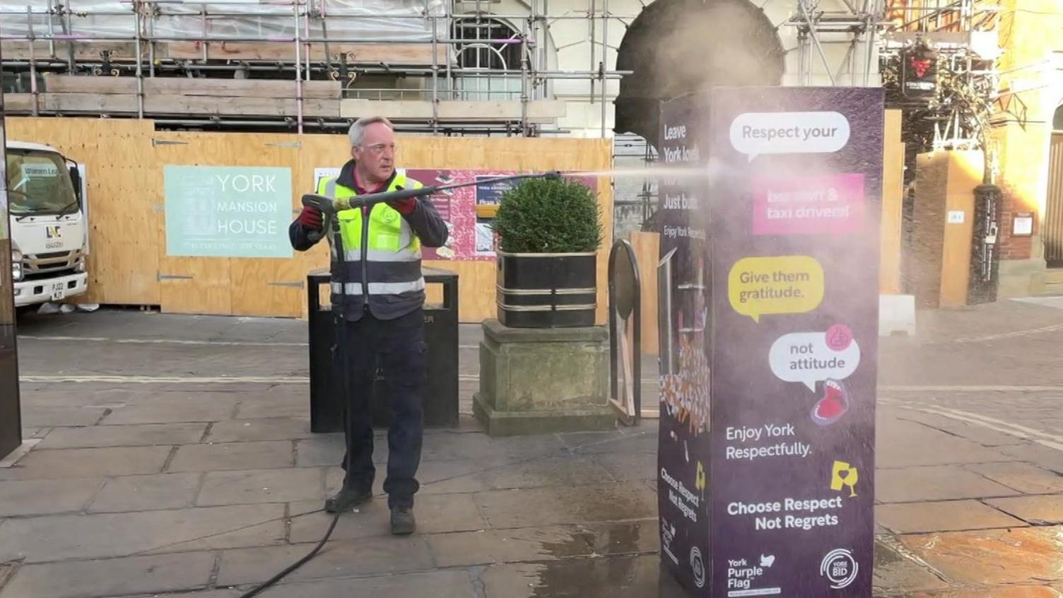 A man in a hi-vis vest jet-washes street furniture in York city centre.
