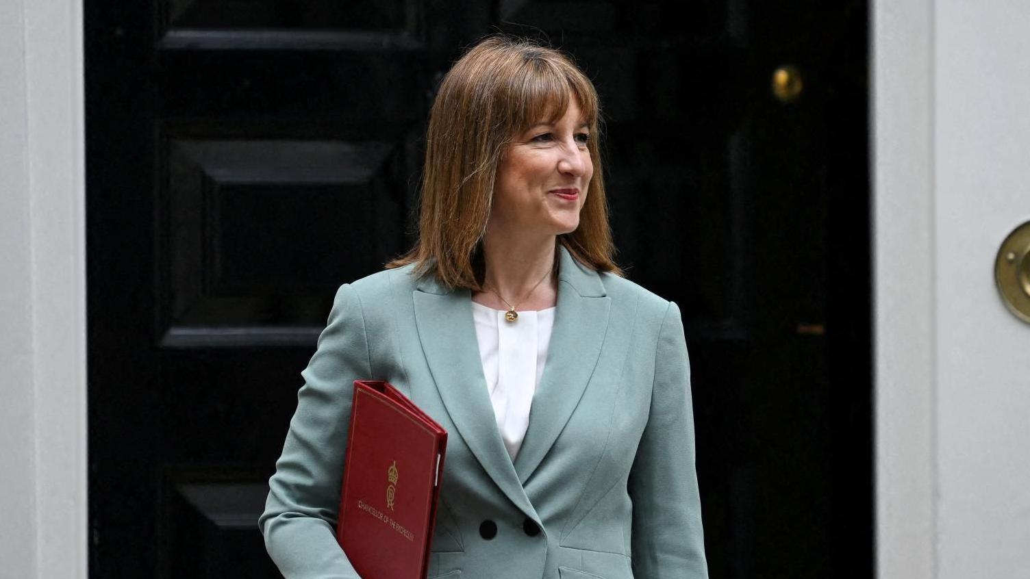 Rachel Reeves wears a light blue suit and is holding a red folder. She is standing in front of the black door of 11 Downing Street.