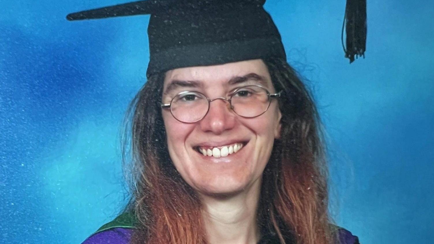 A woman with brown hair and round glasses is smiling in a graduation photo.  She is wearing a cap and gown.