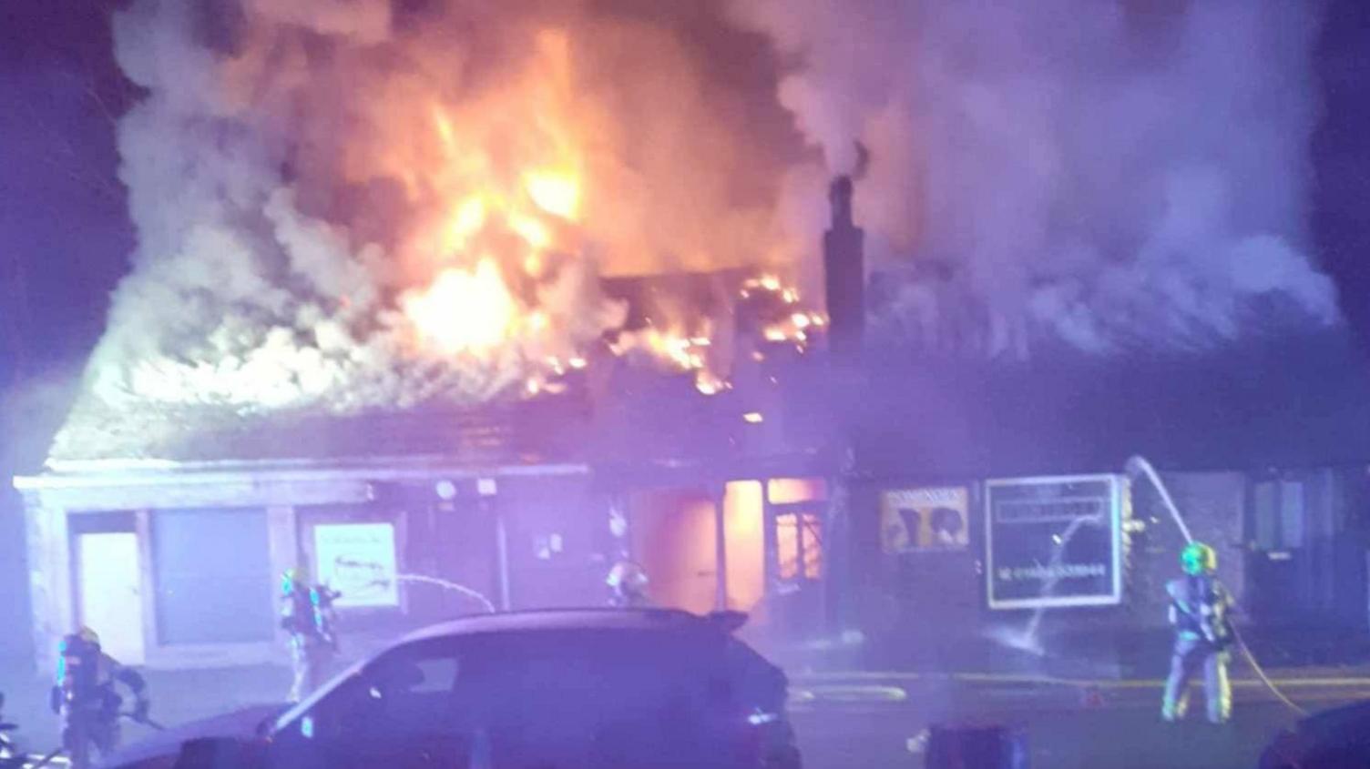 A row of single storey buildings on fire. Flames are coming from the roof, with firefighters using hoses to stem the blaze. 