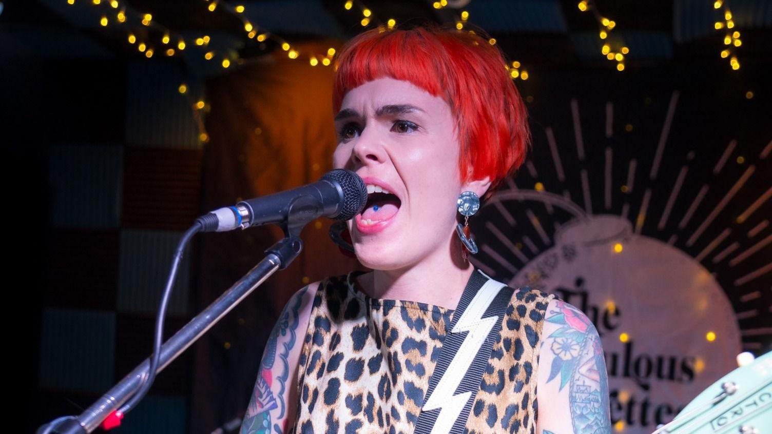 A woman with short red hair sings into a microphone on stage. She is wearing a leopard print dress with a lightning-print guitar strap. She has many tattoos. 