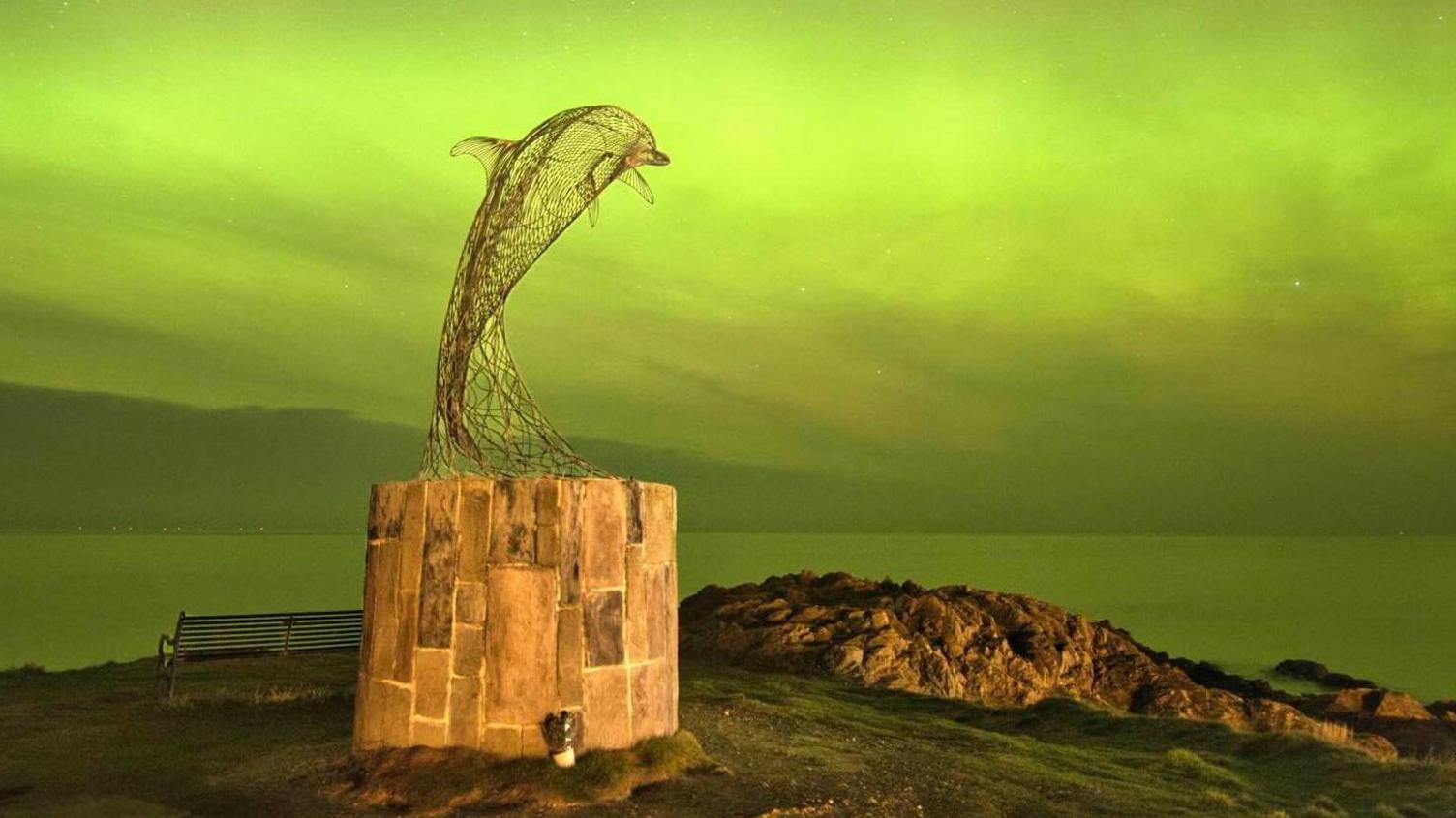 A statue of a dolphin against a backdrop of bright green sky over water.