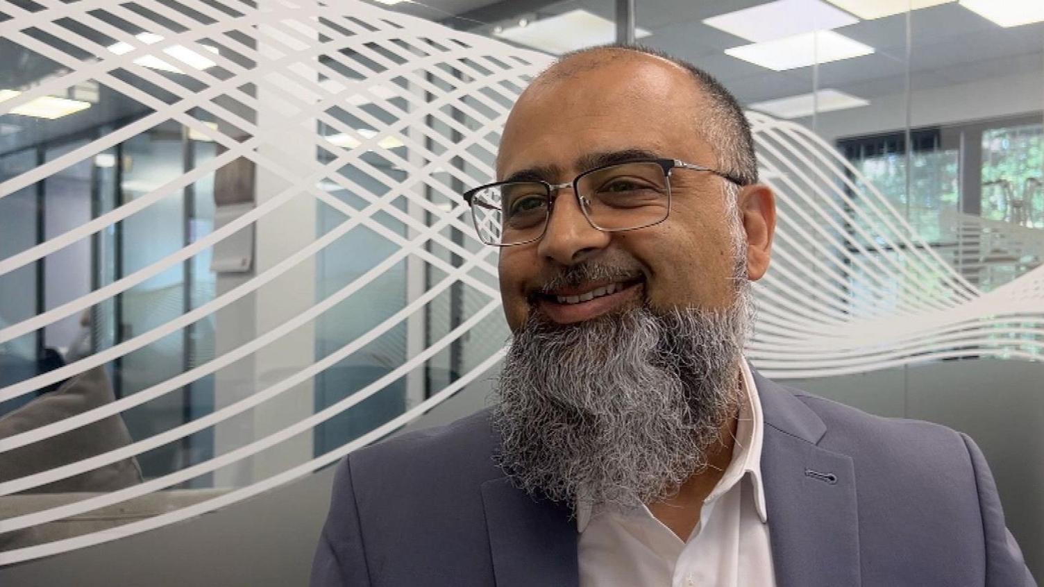 Dewsbury and Batley MP Iqbal Mohamed sits in his constituency office with a pale blue suit on. He is smiling and has a greying beard and glasses.