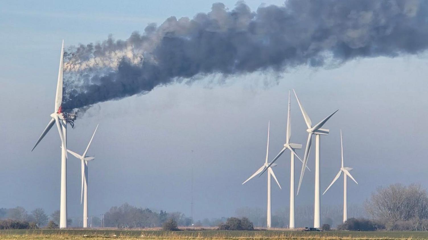 Smoke is billowing from a wind turbine that is on fire in a field. There are five other turbines nearby. The sky is blue and there is a field in the foreground and trees in the background