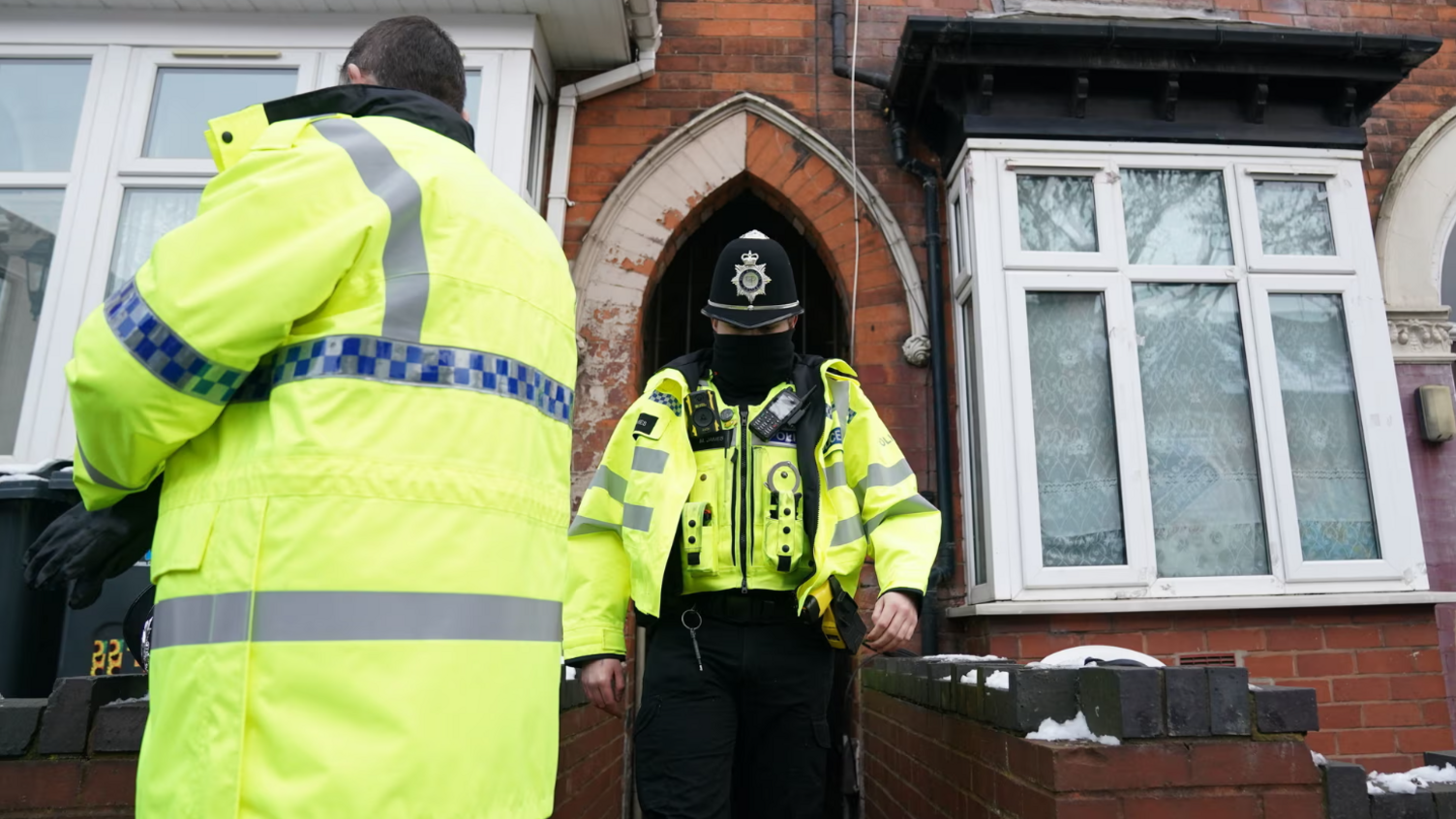 Two police officers leaving a red brick house with white windows. Both officers are wearing fluorescent jackets, with one in a police had and a facemask