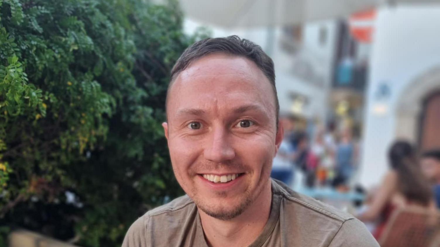 Dan Prescott, who has short brown hair and is wearing a grey t-shirt, smiling directly into the camera. The background is out of focus. There is a tree behind him and - further in the distance - what could be a shopping centre.