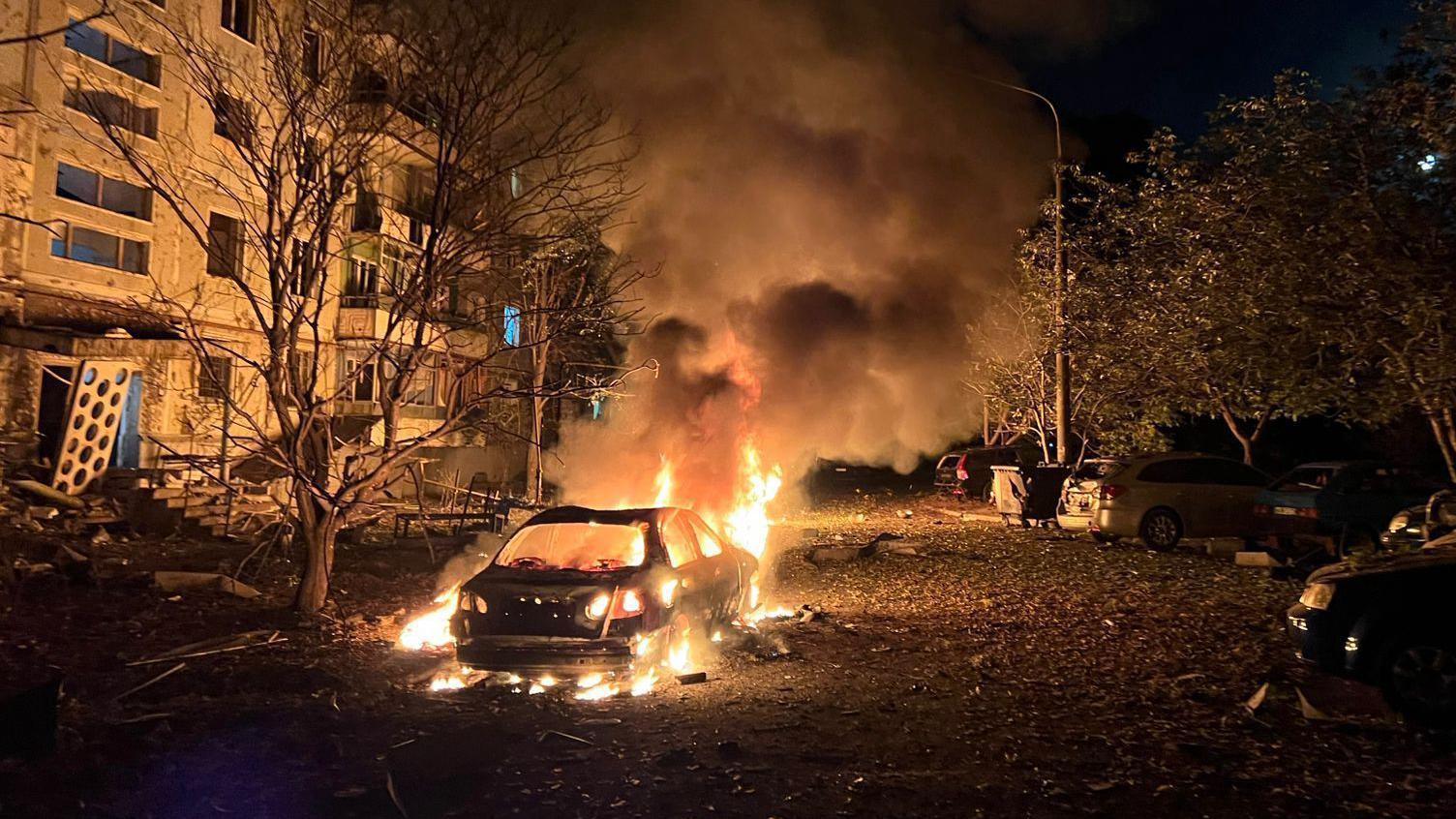 Flames rising from a car on fire in front on an apartment building, during the night.