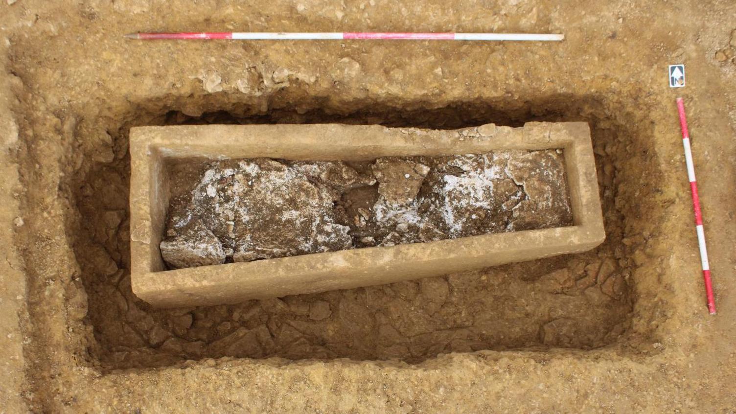 Bird's eye view of an stone coffin, with no lid, still in the ground, but with earth removed down to the bottom of the sarcophagus, revealing its shape