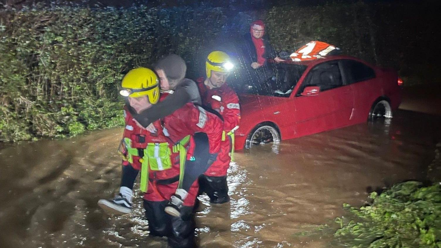 Two firefighters are carrying Ashley Stevens through flood water away from the car to safety. Davie Woodley-Kingston is still sitting on his car behind them.