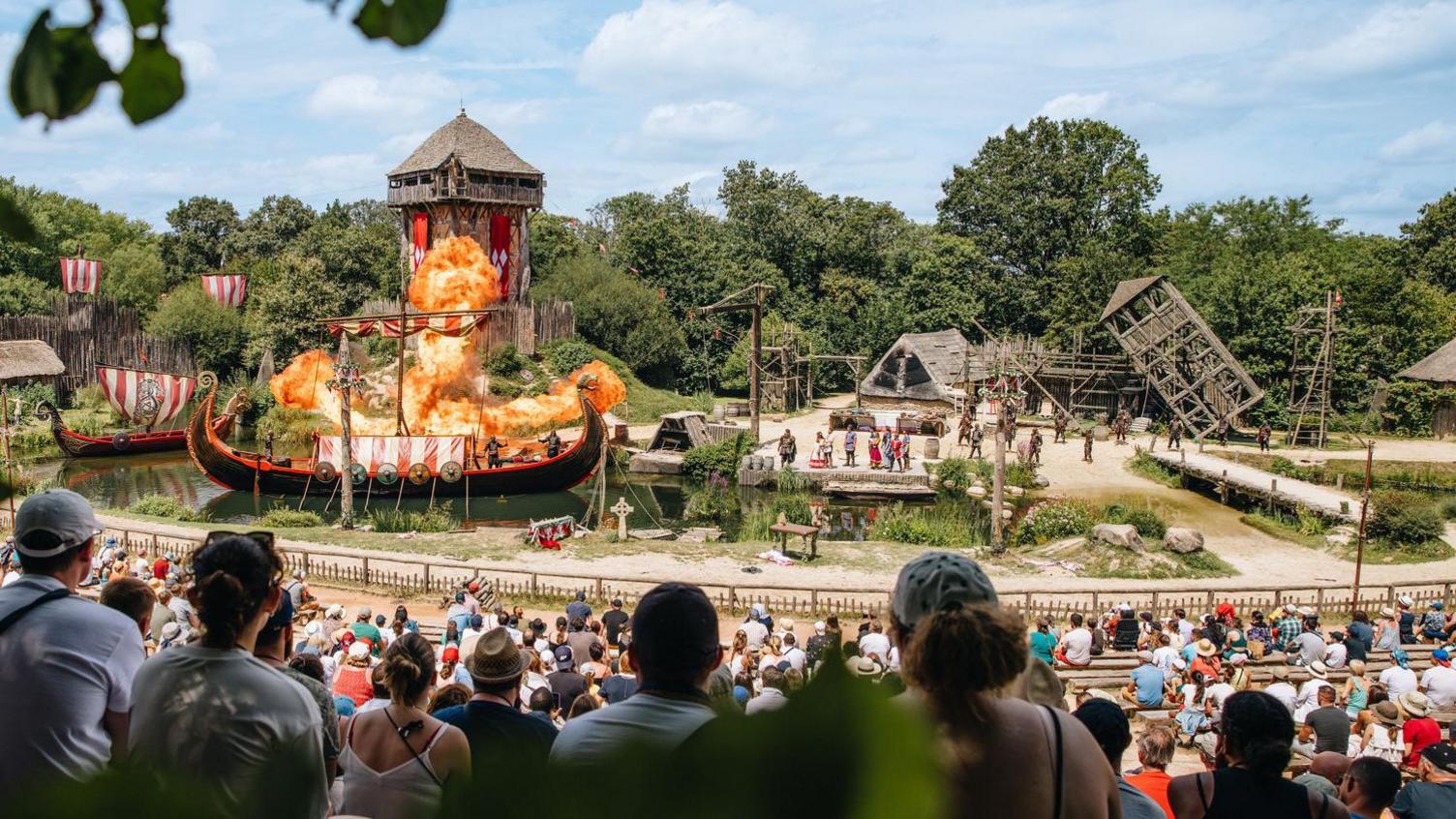 A crowd sat watching a large Viking based show at a Puy du Fou park. The scene is centred on a large Viking longboat which appears to be on fire.