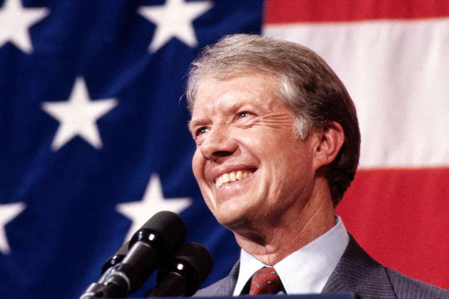 Jimmy Carter smiles in front of a microphone with American flag in the background, at a town meeting in Elk City, Oklahoma 1979.