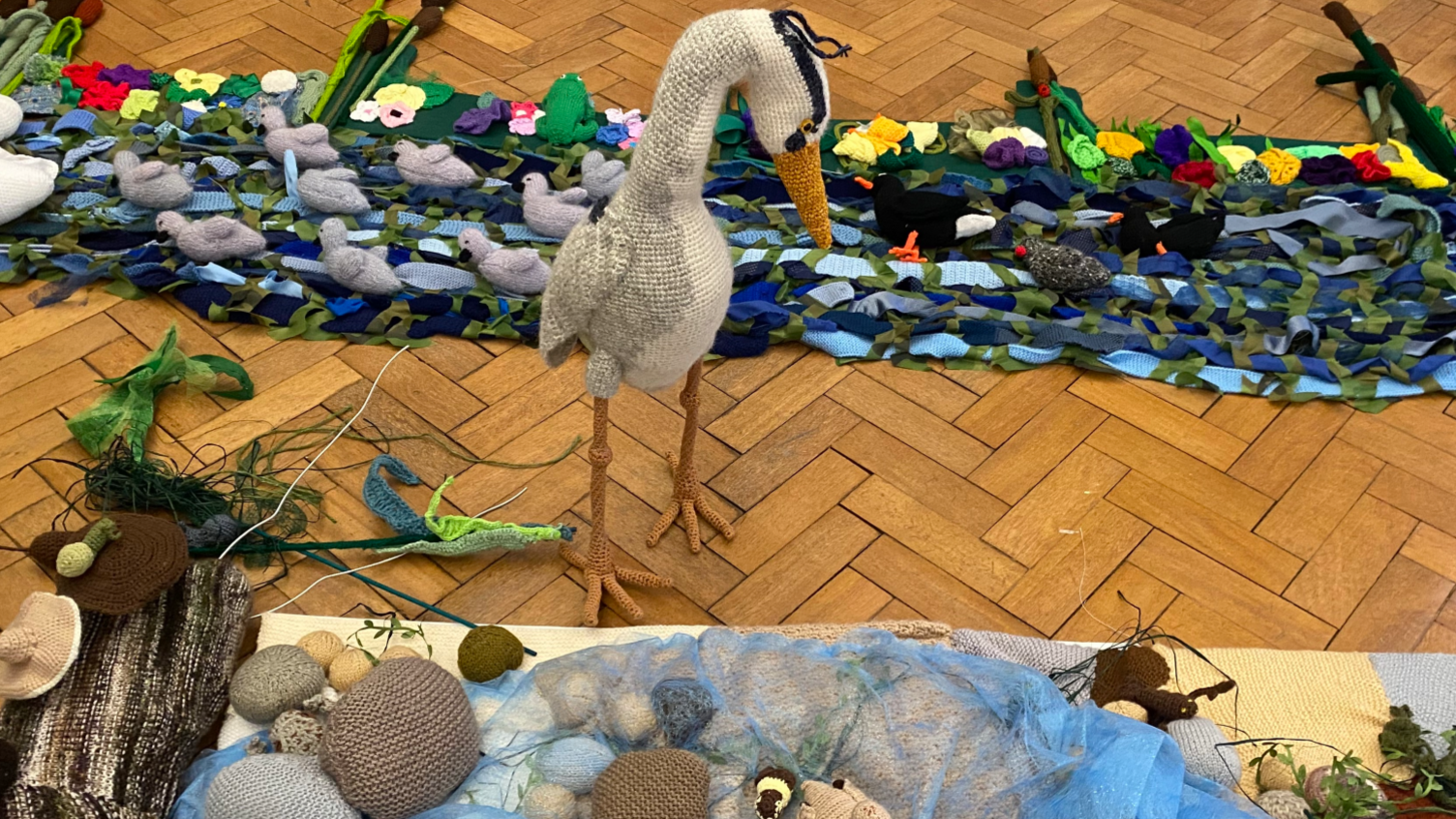 A heron, looking at a river scene, all made out of fabric, the heron is standing up. 