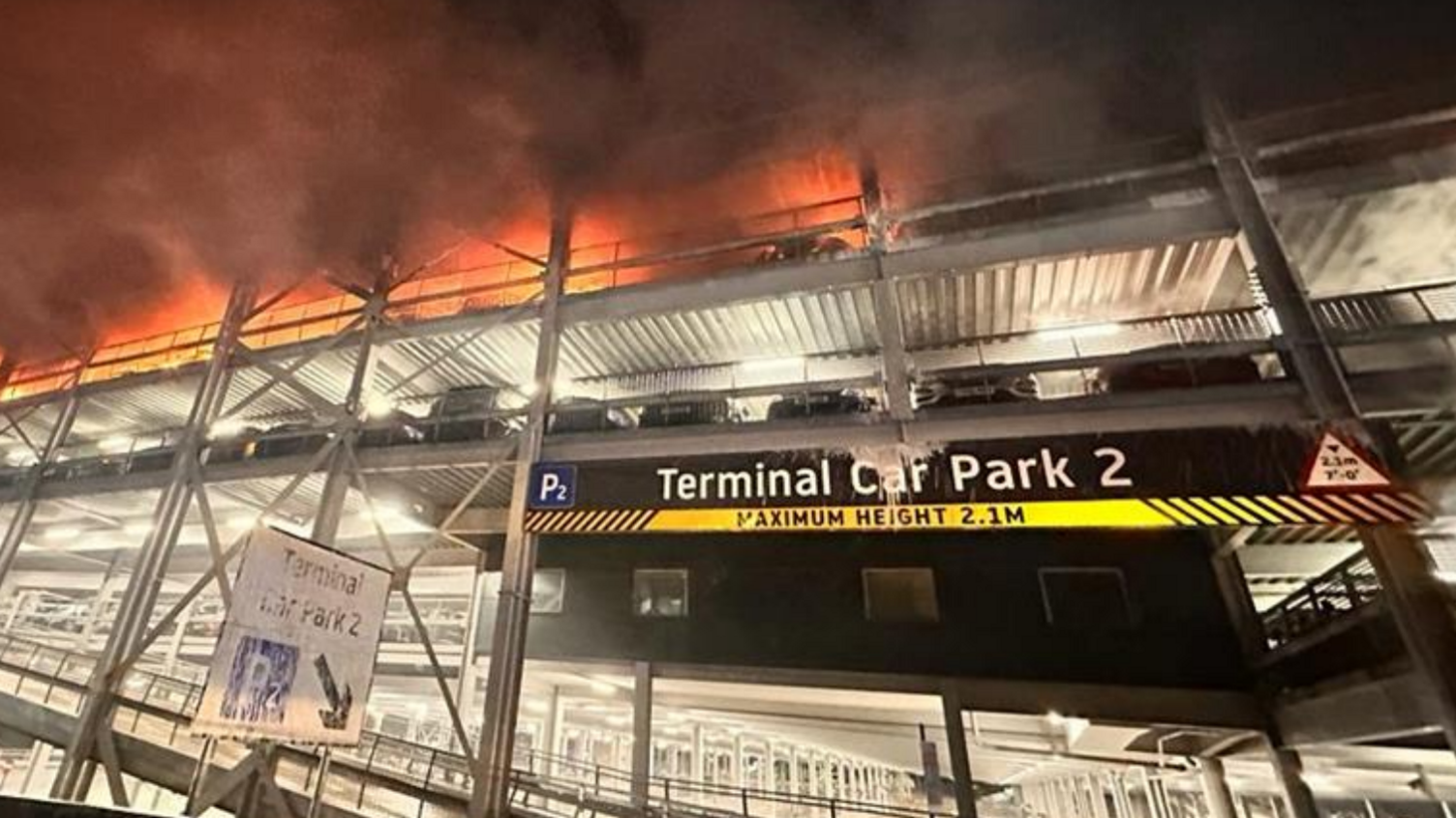 The third floor of Car Park 2 is on fire. Cars can be seen parked on the floor below. The floors above are obstructed by thick black smoke.