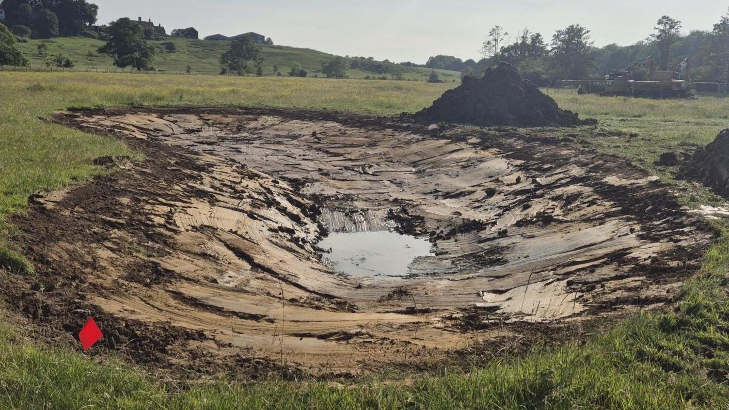 A pond that has been dug, showing water in the middle, grass around it, a mound, trees in the distance and machinery to the right. 