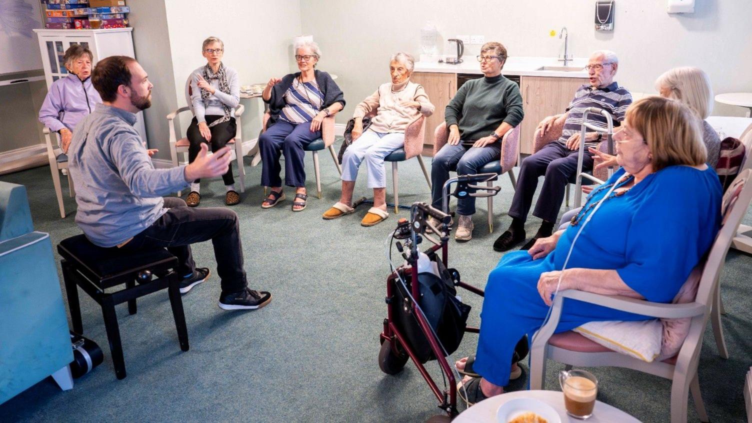A group of elderly people sat on chairs, attentively listening to a man who is gesturing to them