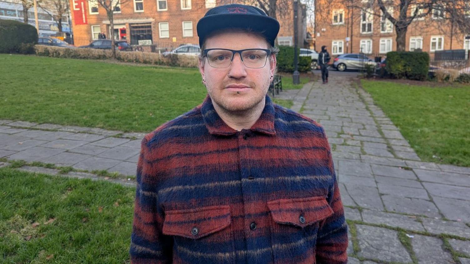 Simon Roche wearing a cap and glasses standing on a green space outside in Bristol looking at the camera