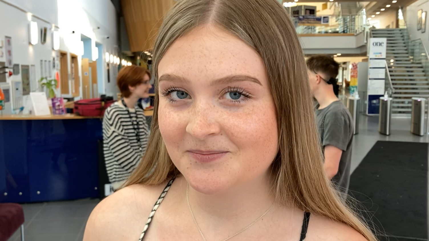 Megan Green. who has long brown hair and freckles, smiles at the camera in the college foyer