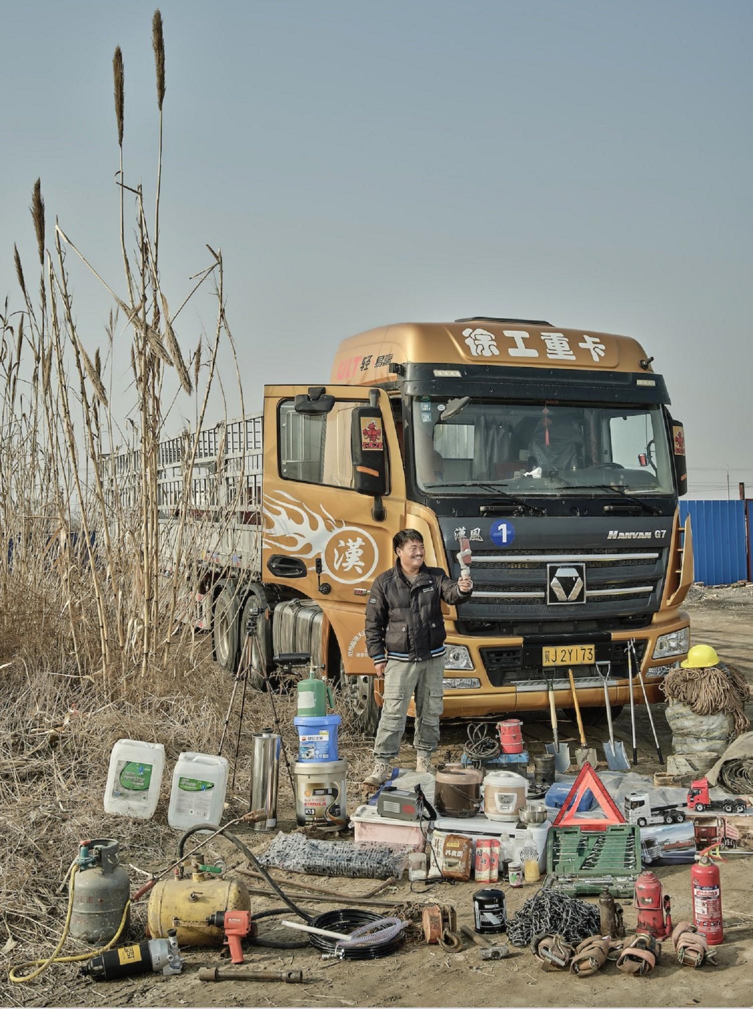 A man takes a selfie while standing in front of his truck, with trucking equipment and supplies laid out in front of it