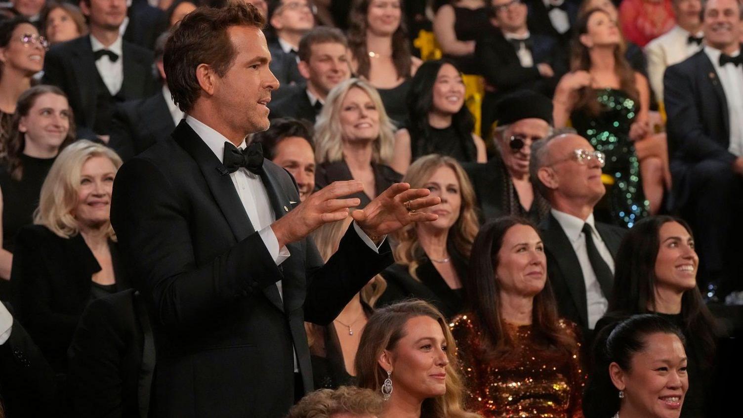 Ryan Reynolds wearing a tuxedo stands with his hands in front of him and his wife Blake Lively sits with a straight smile 