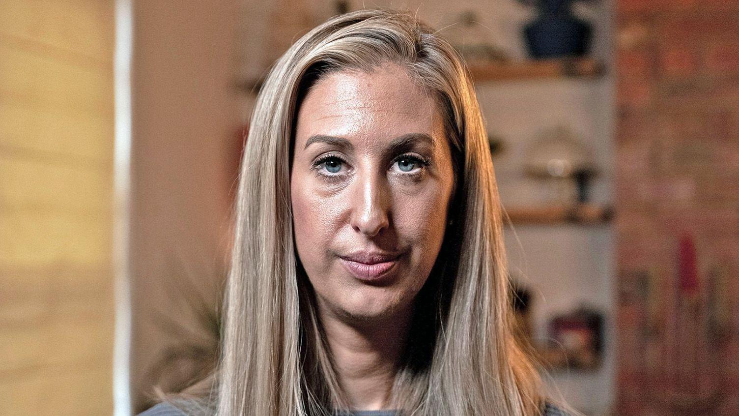 Portrait photo of Fiona Winser-Ramm. She has long, straight blonde hair with a side parting. She has blue eyes and is wearing a grey ribbed jumper. She is pictured in a living room, with shelves and a blind visible behind her.