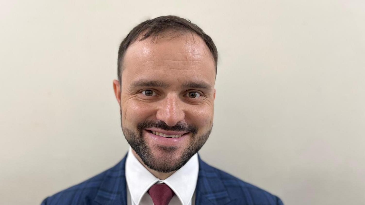 A man with short dark hair and a trimmed beard wearing a white shirt, blue and black suit jacket and dark burgundy tie