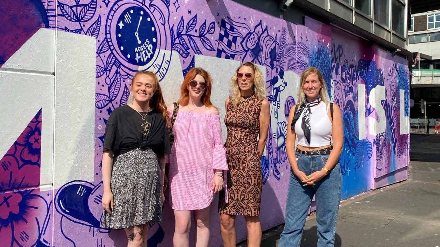 Four women stand in front of a lilac mural painted on a wall, which has dark blue and dark purple features, and white lettering on top.