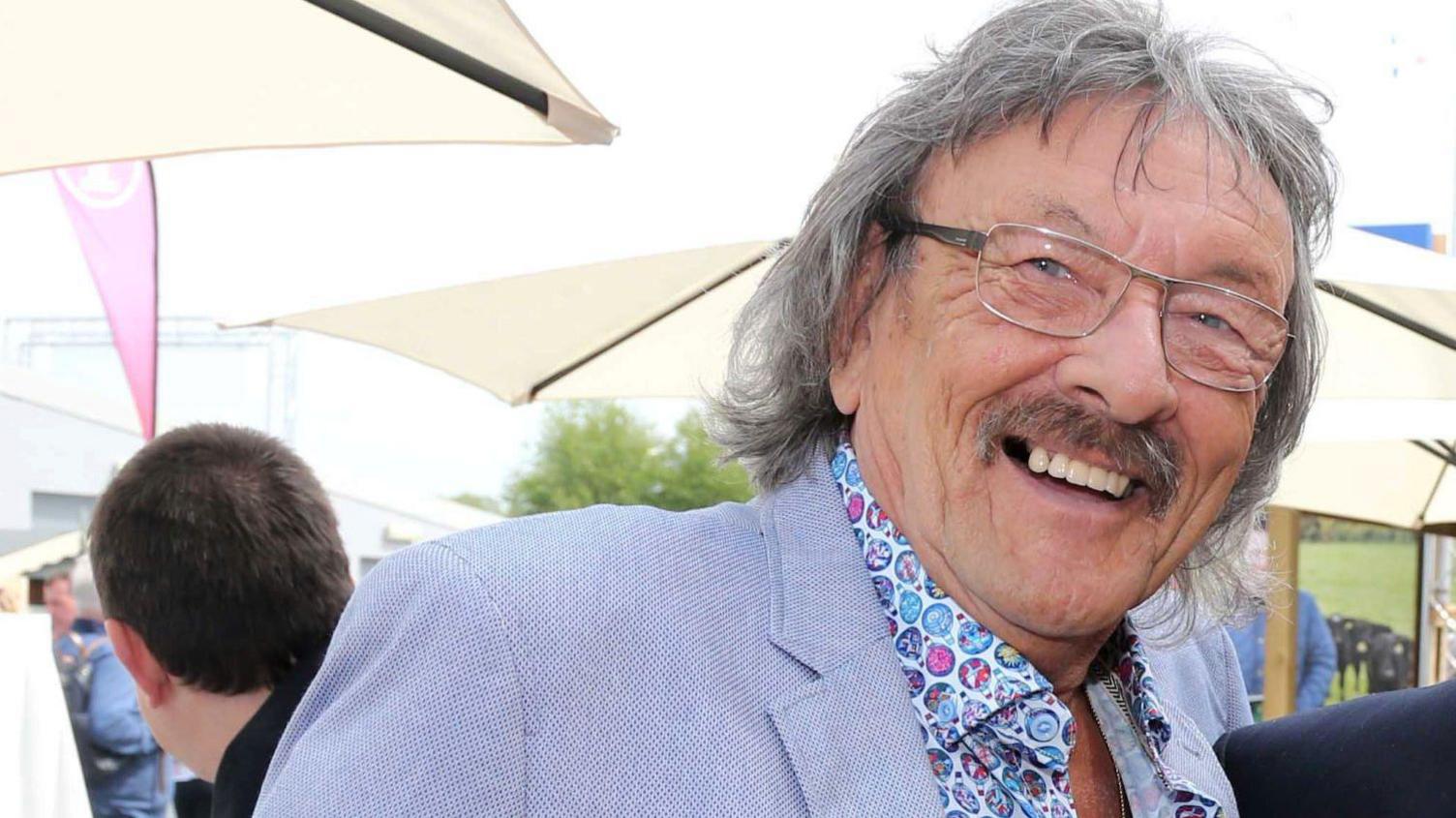 Trevor Campbell smiles at the camera. He has long grey hair and a grey moustache. He has on glasses and a blue blazer with blue multi-coloured shirt. Behind him are cream umbrellas.