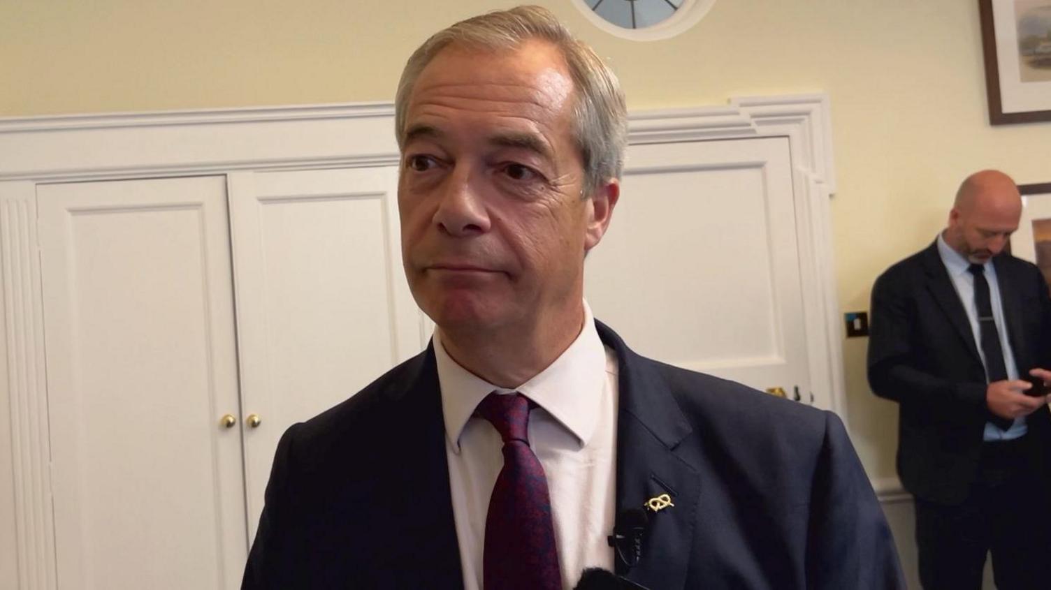 A man looking forwards and to the side of the camera. He is wearing a dark suit and dark purple tie and white shirt. Behind him are white cupboards and another man who is wearing a suit is looking down.