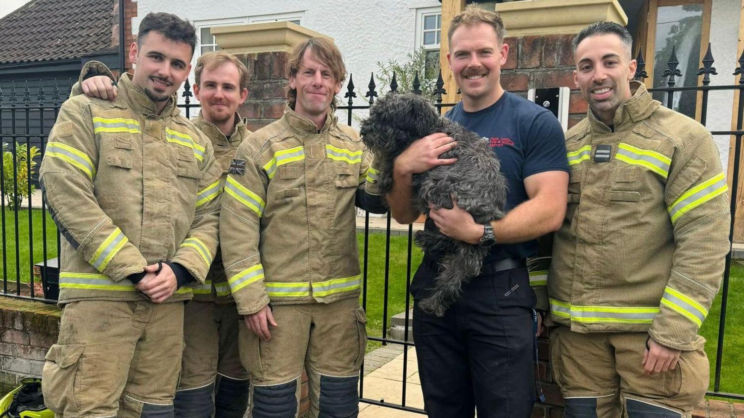 Five firefighters wearing their khaki and yellow uniform huddled together outside the black metal garden fence. One firefighter is wearing a navy T-shirt and has a moustache.