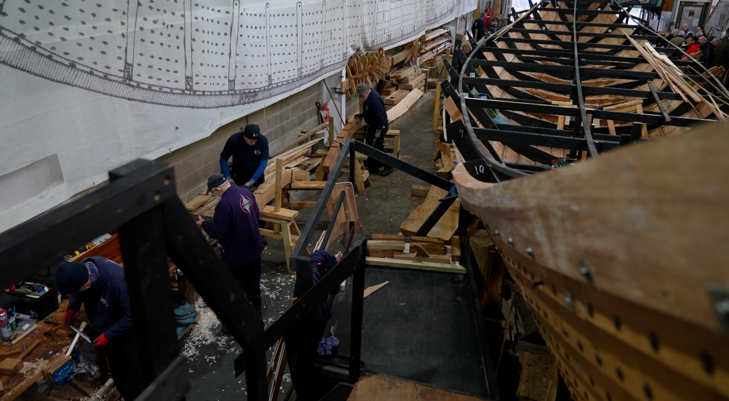 An aerial view of a long wooden boat being worked on by three men. The men are all dressed in dark blue clothing and scraps of wood are on the floor of their workshop. 